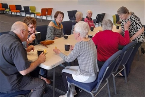 a group of older people sit around a table drinking hot drinks and talking